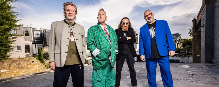 Four performers in colourful blazers stand together outdoors against an urban backdrop.