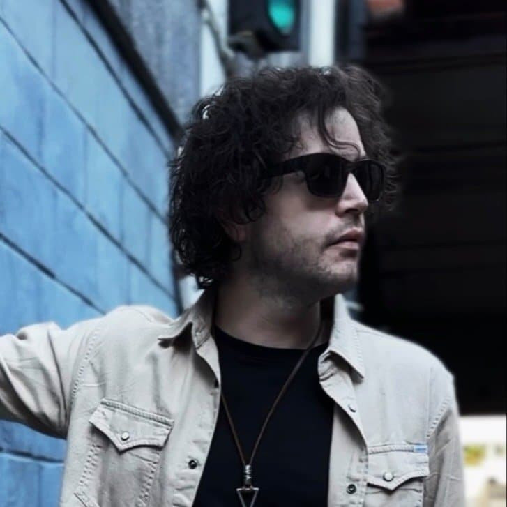 Man with dark hair and sunglasses wearing a cream denim jacket over black shirt against an urban blue wall.
