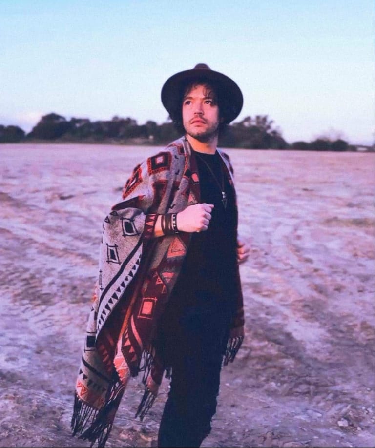 Man in black hat and shirt wearing patterned shawl stands in open field at dusk.