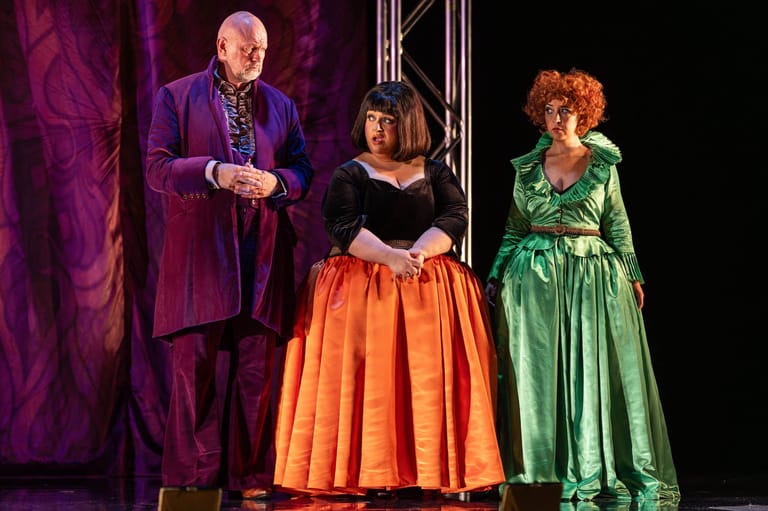 Three actors in period costume stand on stage under purple lighting, with a woman in an orange skirt centre stage.