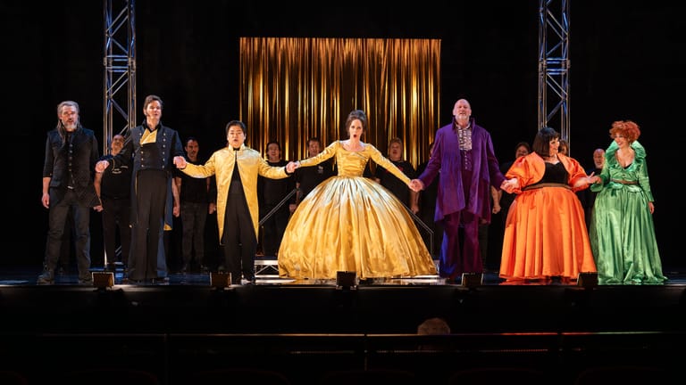 Cinderella in a gold ball gown stands centre stage with fairy tale characters in jewel-toned costumes flanking her beneath golden curtains.