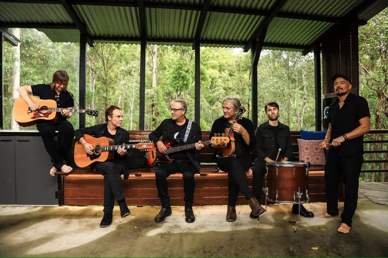 Six musicians in black clothing perform with acoustic guitars and percussion in a glass pavilion surrounded by trees.