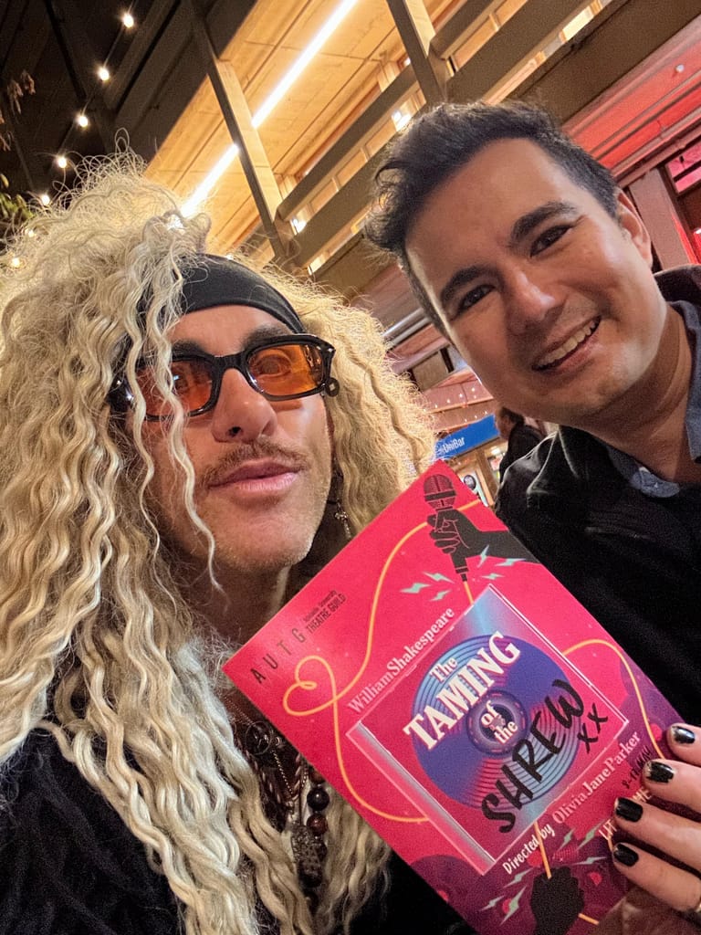 Two people smile at the camera while holding a bright pink programme for The Taming of the Shrew inside a theatre foyer.