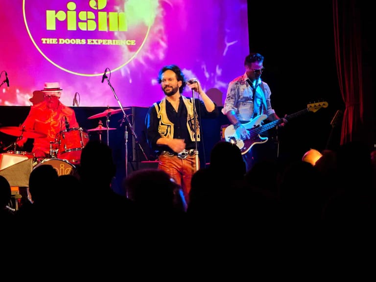 Three musicians perform on stage under purple and pink stage lighting during The Doors Experience tribute show.