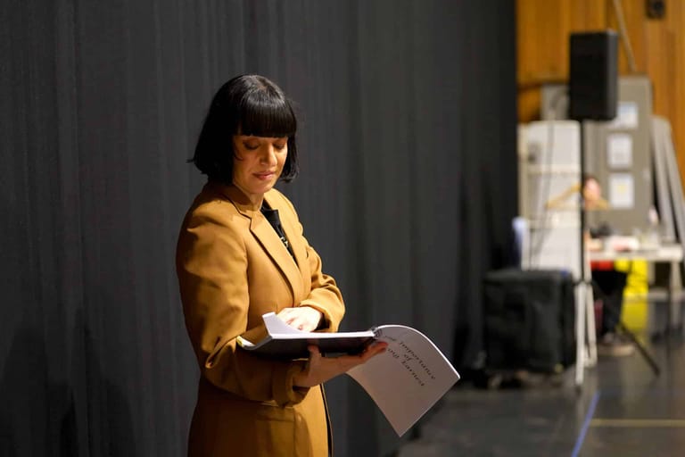 Woman in mustard blazer holding papers stands against grey curtain backdrop during interview.