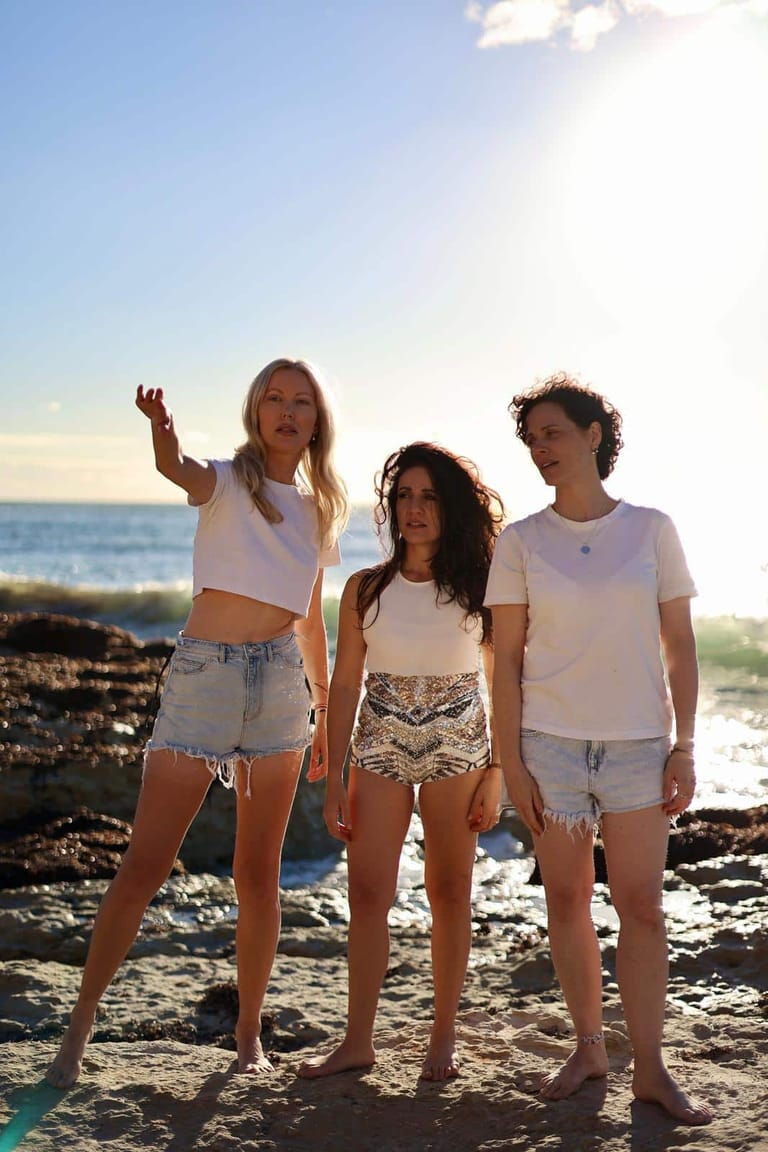 Three young women in white tops stand together on a rocky beach at sunset.