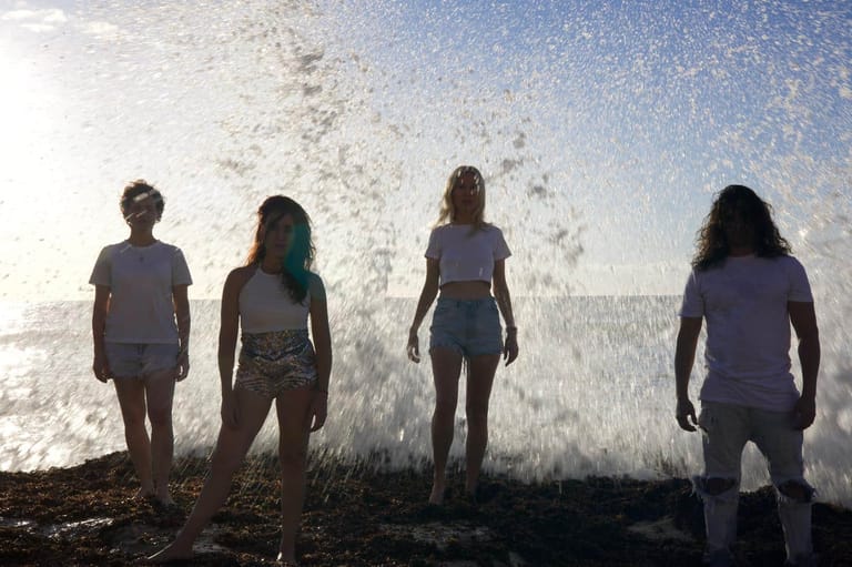 Four performers stand silhouetted against crashing ocean waves during an outdoor theatre production.