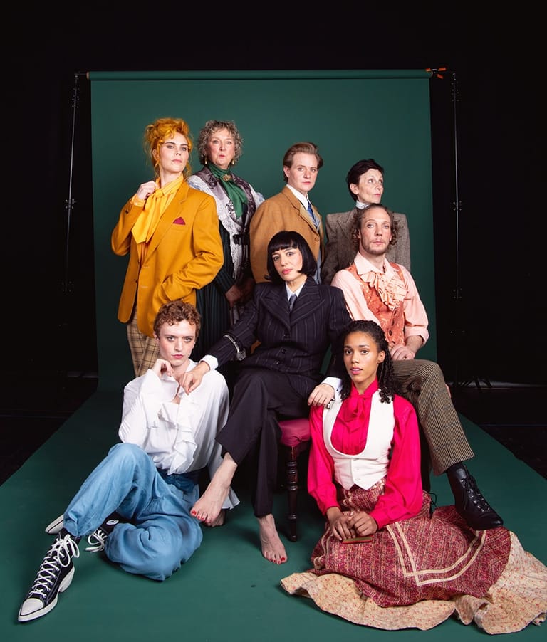 Eight actors in period and contemporary costume pose on a green stage set for a theatrical production.