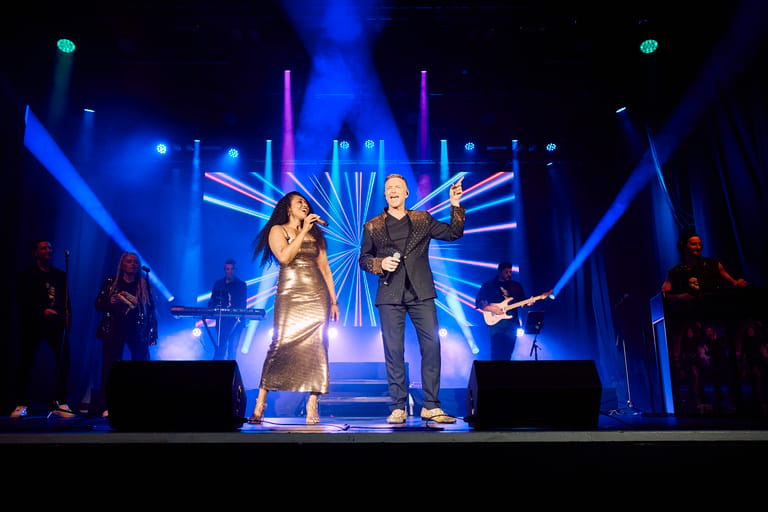 Two performers stand centre stage beneath blue and purple stage lighting with geometric set design behind them.