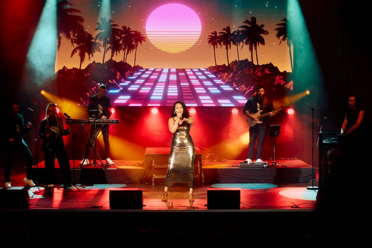 Singer in white dress performs on stage with neon sunset backdrop and palm tree silhouettes.