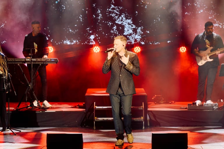 Band performing on stage with lead vocalist centre stage, surrounded by red lighting and pyrotechnics.