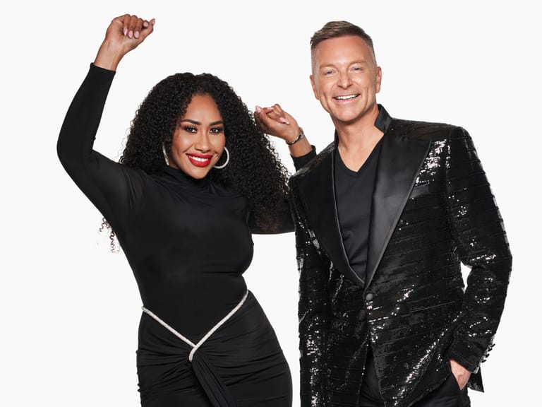 Two people in sequined black outfits pose together with raised fists against a white background.