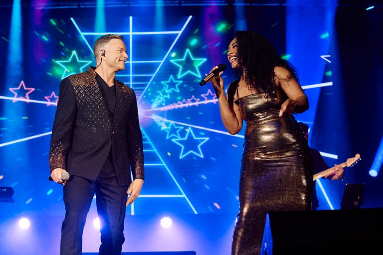 Two performers on stage with microphones against a vibrant blue neon star-patterned backdrop.