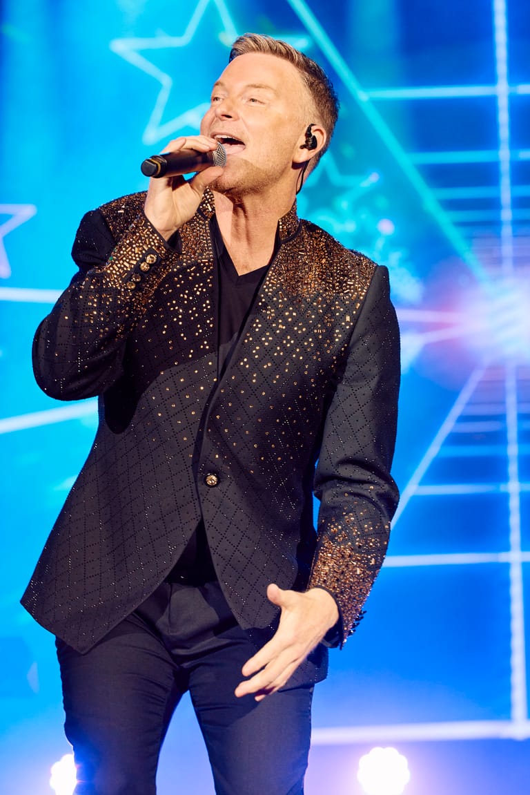 Performer in a sequinned blazer singing into a microphone against a vibrant blue neon stage backdrop.