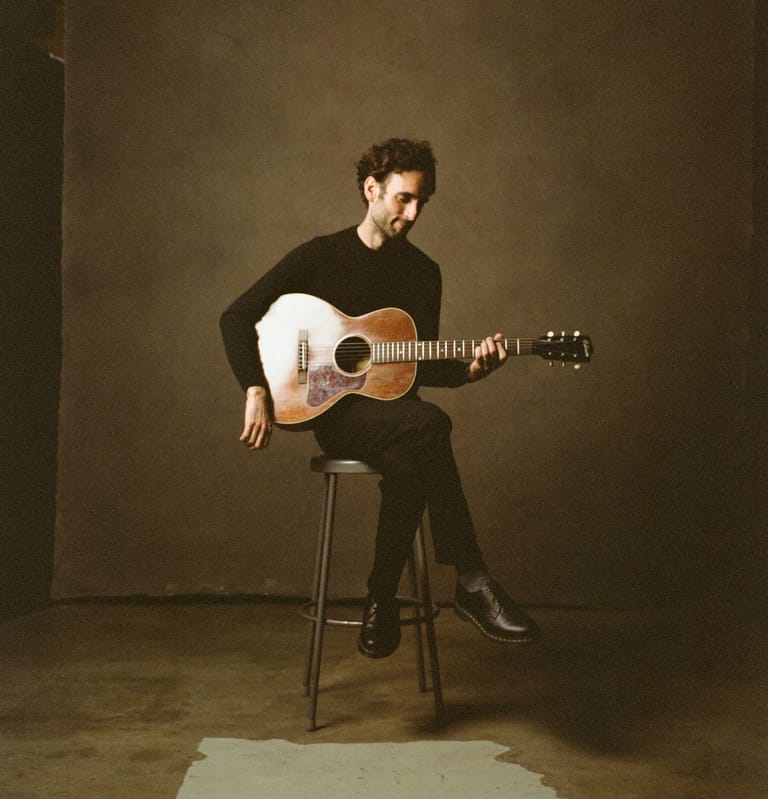 Musician seated on a stool playing an acoustic guitar against a warm brown backdrop.