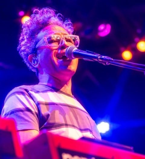 Performer wearing glasses singing into a microphone at a keyboard, bathed in vibrant purple and pink stage lighting.