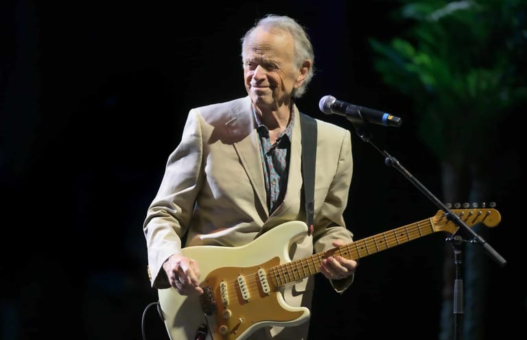 Musician in cream blazer playing electric guitar at microphone on stage.