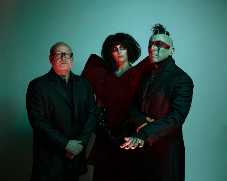 Three musicians pose against a teal background, dressed in dark clothing with distinctive styling and accessories.