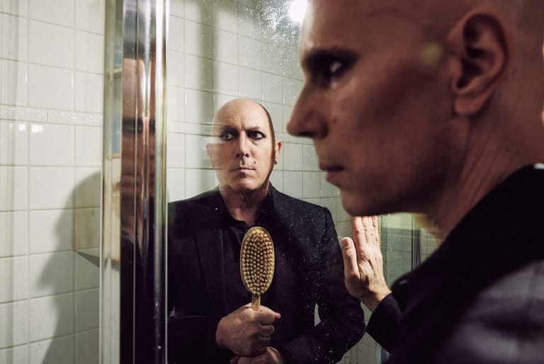 Bald man in glasses holding a hairbrush, examining his reflection in a bathroom mirror.