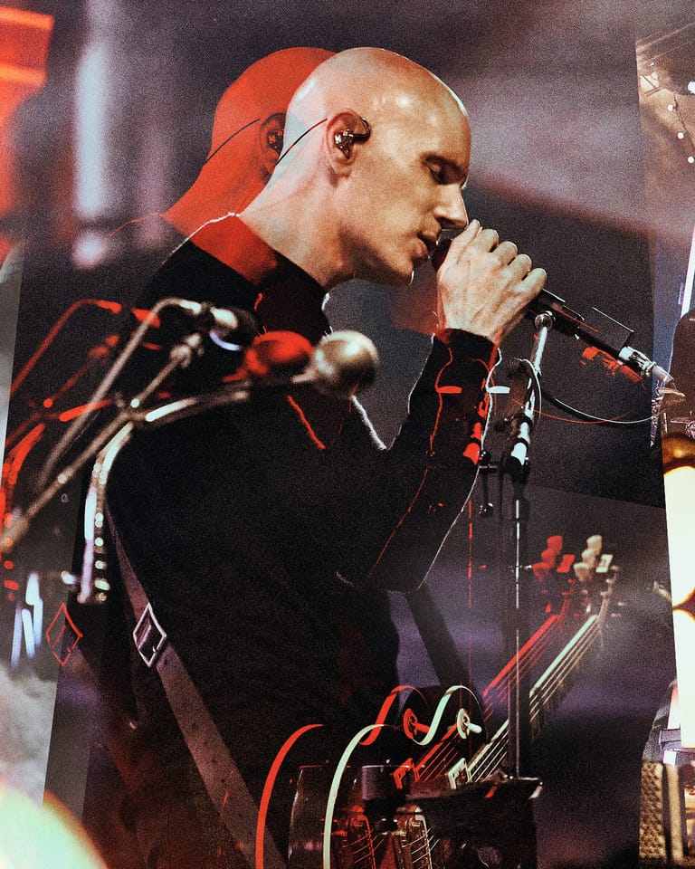 Bald performer singing into a microphone on stage, wearing a red and black patterned jacket under warm stage lighting.
