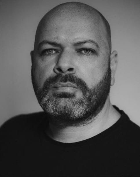 Man with shaved head and full beard in black-and-white portrait photograph.