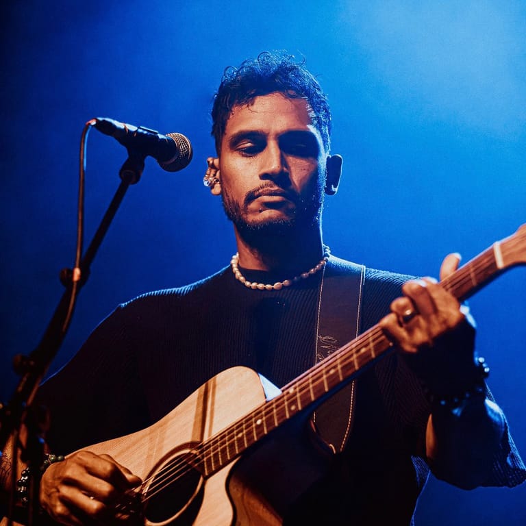 Musician performing with acoustic guitar at microphone against blue stage lighting.