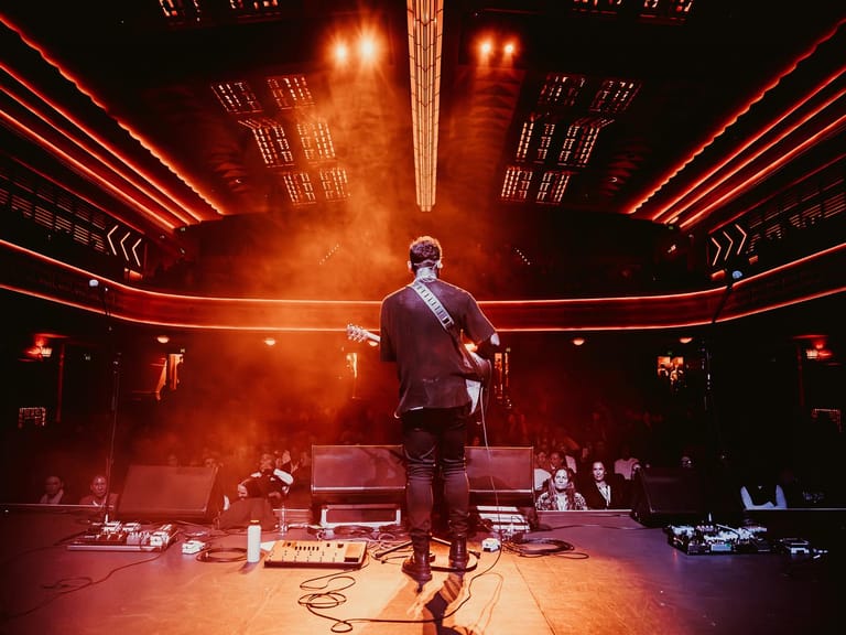 Performer stands centre stage in burgundy suit under dramatic red stage lighting and geometric ceiling arrays.