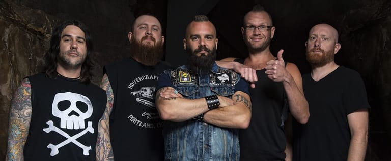 Five bearded band members in dark clothing pose together against a black background.