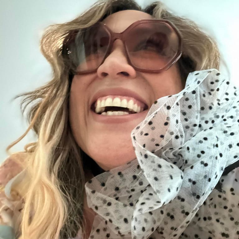 Person wearing oversized rose-tinted sunglasses and a polka-dot collar smiles broadly against a white background.