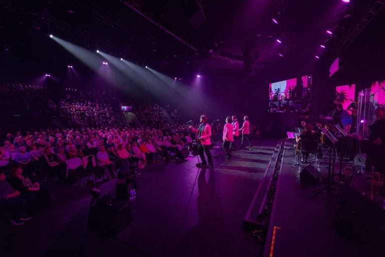 Large crowd seated in an arena watching performers on a stage bathed in purple and pink lighting.