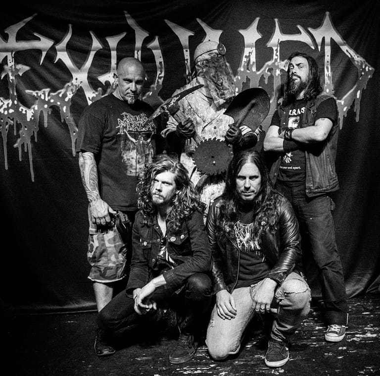 Five metal musicians in band merchandise and studded clothing pose against a dark backdrop with band logo.
