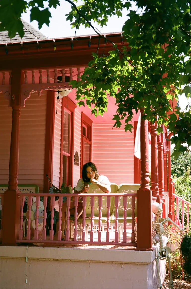 Person seated on the wraparound veranda of a red heritage cottage, dappled by vine shade.