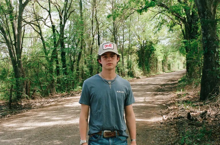 Person in a grey t-shirt and cap standing on a tree-lined dirt path.
