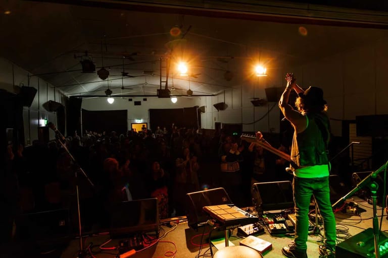 Performer on stage under warm spotlights with audience gathered in dimly lit venue during live event.