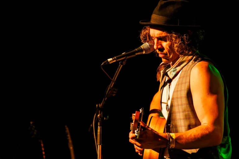 Musician in fedora hat and vest singing into microphone while holding acoustic guitar on stage.