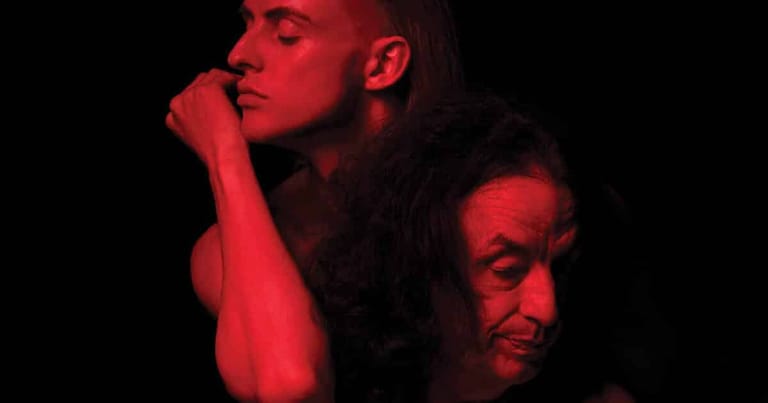 Two figures lit by red stage lighting, one resting chin on hand in contemplative pose.