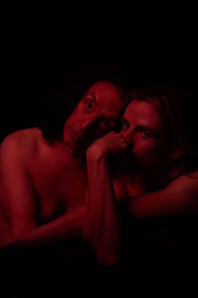 Two figures pose intimately against a black background, bathed in deep red light.