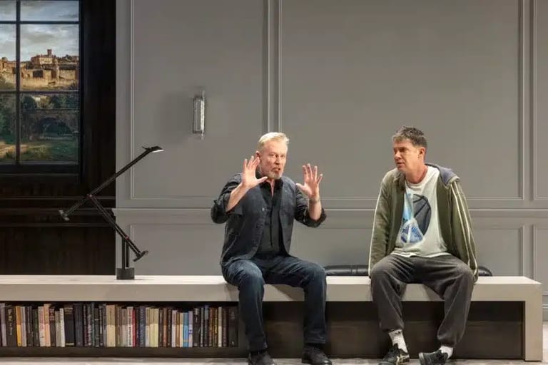 Two actors seated on a white platform engage in animated conversation during a stage production, with bookshelves and windows visible behind them.