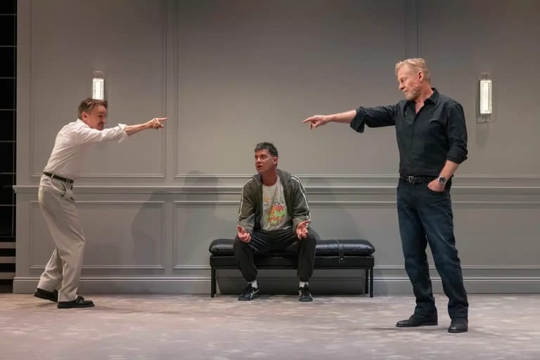 Two men extend their arms toward each other across a stage while a seated figure watches from a black bench between them.