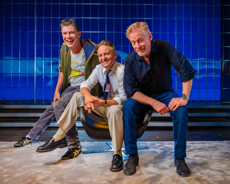 Three male actors crouched together on stage against a bright blue gridded backdrop during a theatrical performance.