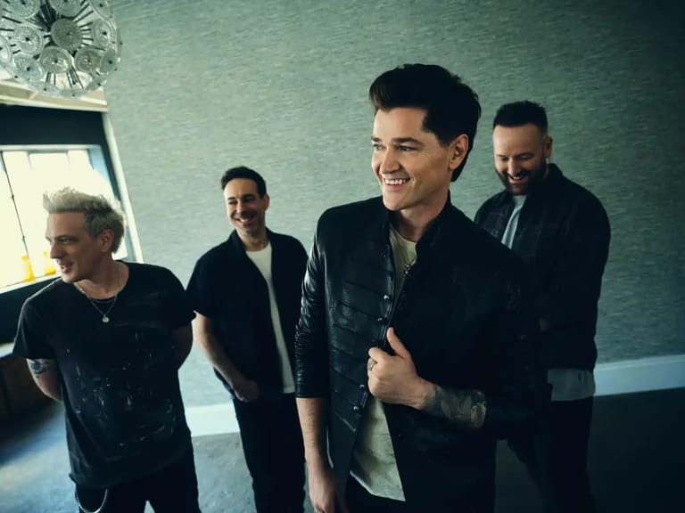 Four musicians in dark clothing stand together in a green-walled room, smiling toward the camera.