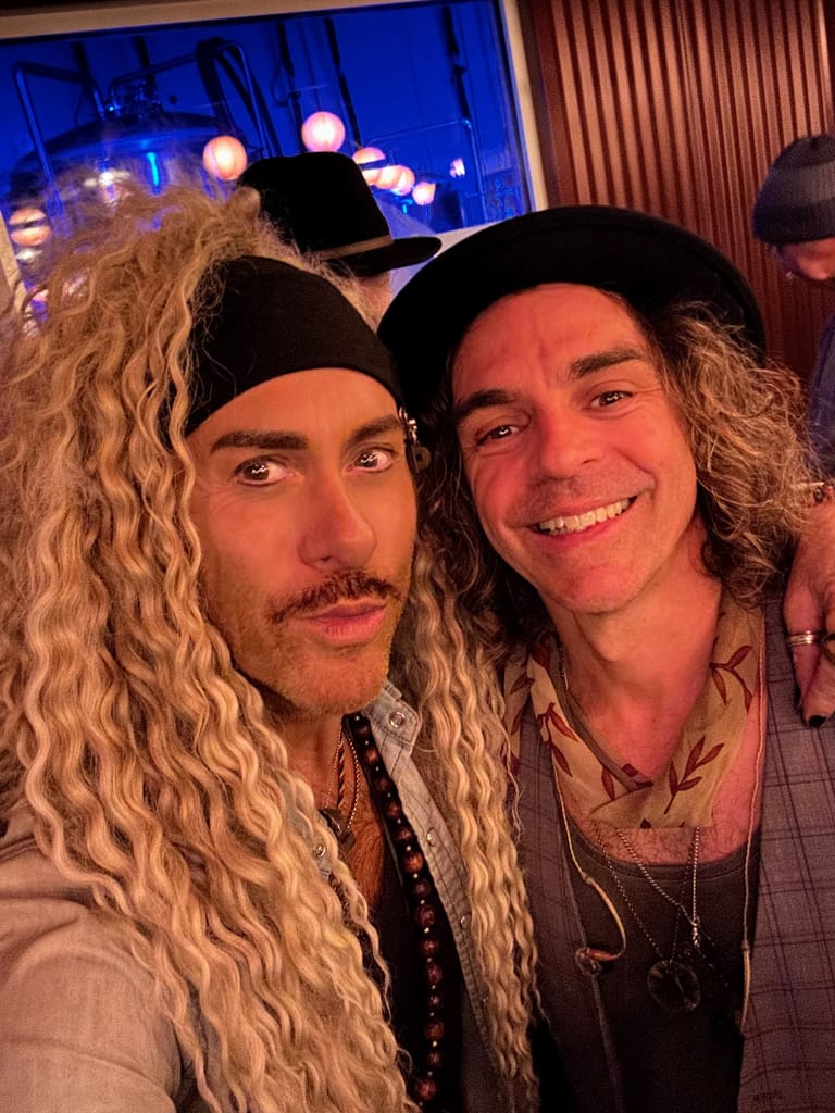 Two musicians with long curly blonde hair pose together at a venue with blue and red stage lighting in the background.