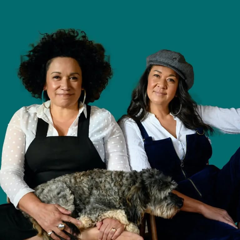 Two women hold a tabby cat against a teal background, both smiling at the camera.