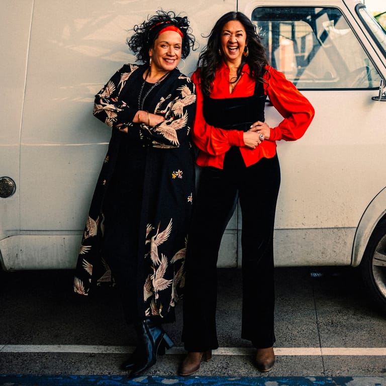 Two people in stylish outfits pose cheerfully beside a vintage white van.