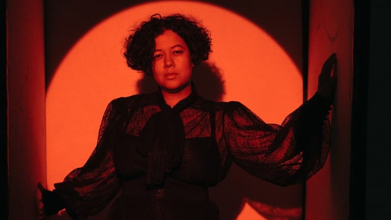 Person in dark clothing posed against a large glowing orange circular backdrop during an interview setting.