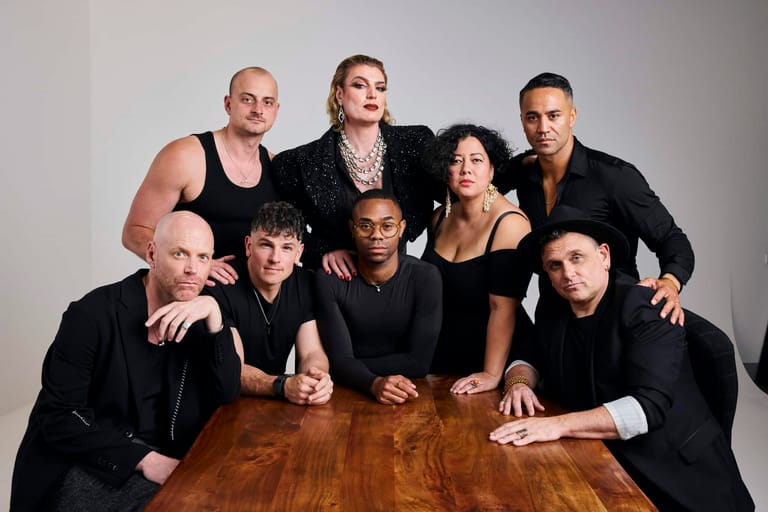 Seven people in black clothing pose together around a wooden table against a white background.