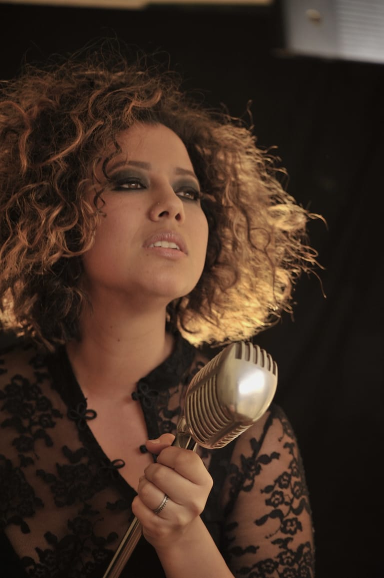 Person with curly auburn hair holding a vintage microphone, gazing upward against a dark background.