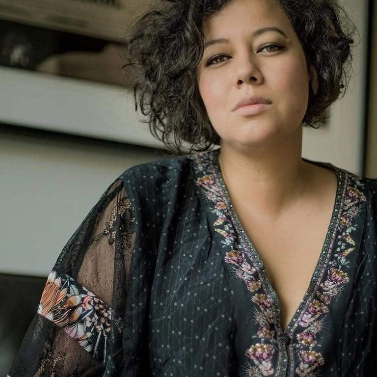 Person with curly dark hair wearing an ornate black patterned robe, gazing directly at camera in an indoor setting.
