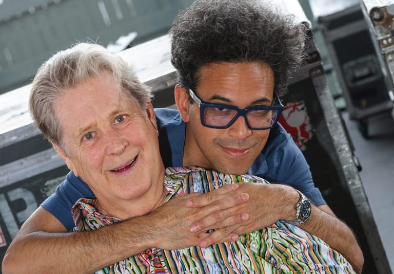 Two people smiling together at an indoor event, one wearing glasses and colourful patterned clothing.