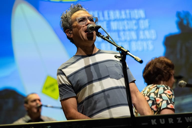 Man in striped shirt speaking into microphone at a panel discussion with blue screen backdrop.